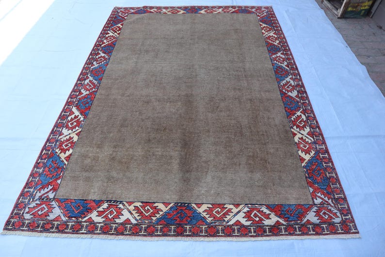 May include: A rectangular, brown rug with a patterned border. The border features a repeating geometric design in red, blue, and white. The rug is likely made of woven material and is suitable for a living room or bedroom.