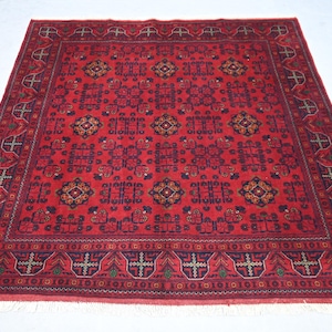 May include: A square, red oriental rug with a detailed geometric pattern. The rug features a central field of repeating shapes in dark blue, orange, and green, framed by a border with similar motifs. The rug has a fringe along the edges.