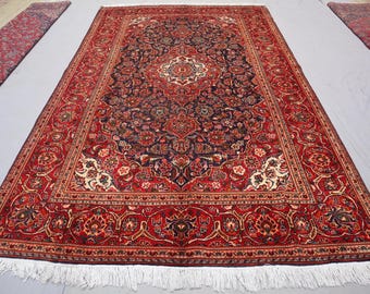 Puede incluir: Una gran alfombra persa rectangular con un diseño de medallón central. La alfombra presenta un borde rojo intenso y un campo azul marino lleno de patrones florales en tonos de rojo, crema y marrón. Flecos blancos bordean la alfombra.