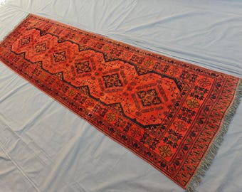 May include: A long rectangular runner rug featuring a vibrant red background with a repeating black geometric diamond pattern. The rug is decorated with intricate black floral motifs and borders, finished with short grey fringe at both ends.