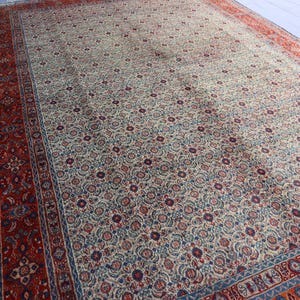 May include: A large rectangular area rug featuring a cream-colored field densely covered in a repeating floral pattern in shades of red, blue, and tan. The rug is framed by a wide, intricate border in deep red with blue and cream floral motifs.