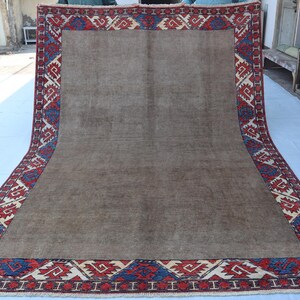May include: A large, rectangular rug with a central brown field and a detailed border. The border features a repeating geometric pattern in red, blue, and cream colors. The rug is displayed on a white surface, showcasing its size and design.