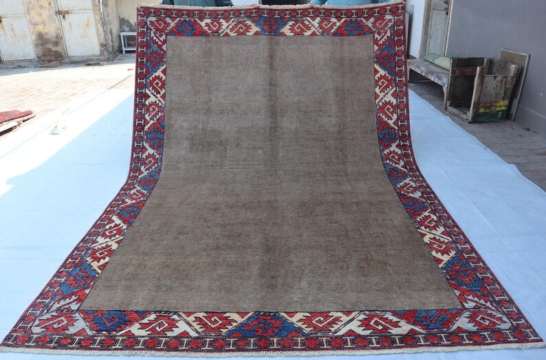 May include: A large, rectangular rug with a central brown field and a detailed border. The border features a repeating geometric pattern in red, blue, and cream colors. The rug is displayed on a white surface, showcasing its size and design.