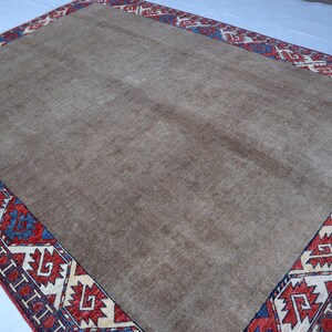 May include: A rectangular, beige rug with a detailed border. The border features a repeating geometric pattern in red, blue, and cream colors. The rug's central field is a solid, neutral beige, providing a simple backdrop to the ornate border.