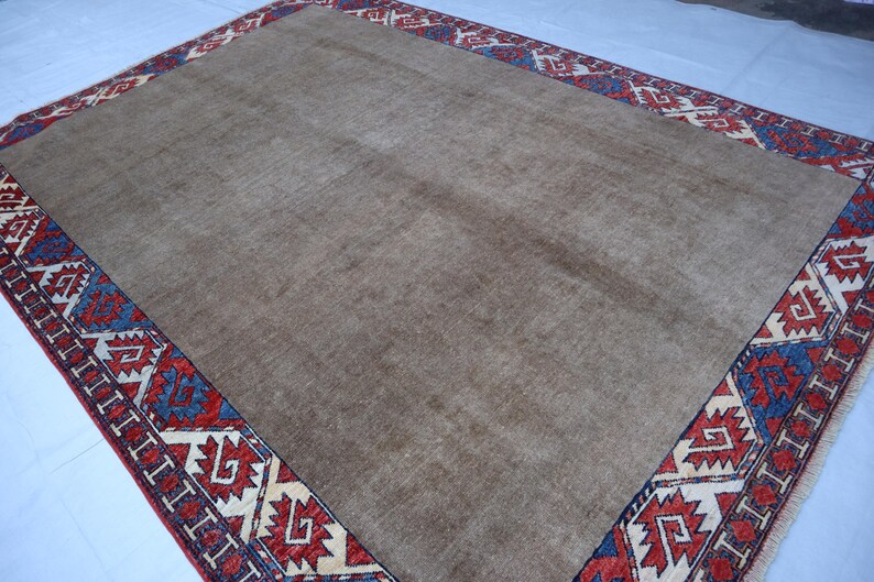 May include: A rectangular, beige rug with a detailed border. The border features a repeating geometric pattern in red, blue, and cream colors. The rug's central field is a solid, neutral beige, providing a simple backdrop to the ornate border.