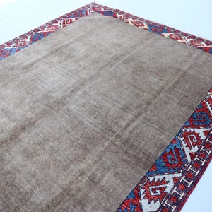 May include: A rectangular, brown rug with a patterned border. The border features a repeating design in red, blue, and white. The rug has a soft, textured appearance, suggesting a comfortable floor covering.