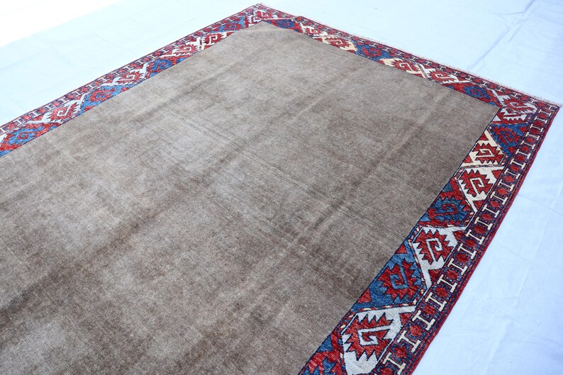 May include: A rectangular, brown rug with a patterned border. The border features a repeating design in red, blue, and white. The rug has a soft, textured appearance, suggesting a comfortable floor covering.