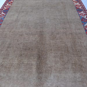 May include: A rectangular, brown rug with a patterned border. The border features a repeating geometric design in red, blue, and cream colors. The rug's central field is a solid, muted brown.