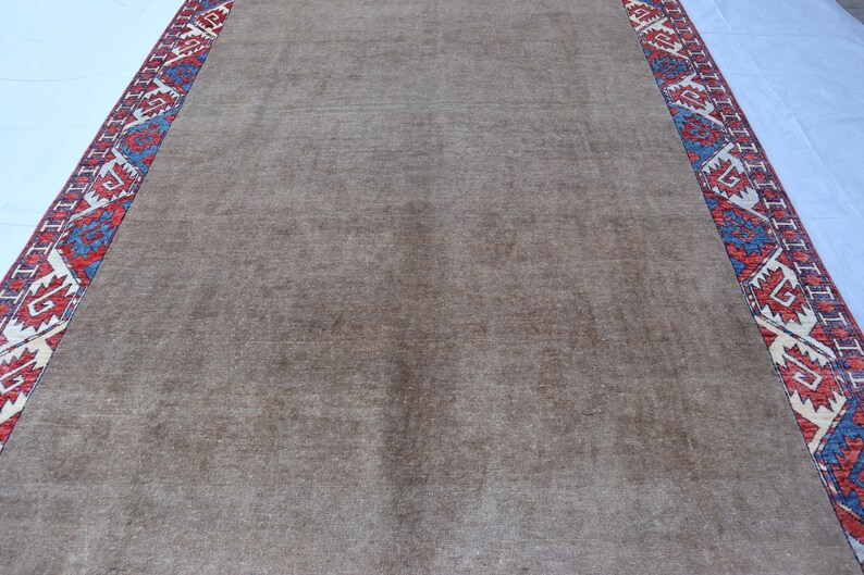 May include: A rectangular, brown rug with a patterned border. The border features a repeating geometric design in red, blue, and cream colors. The rug's central field is a solid, muted brown.
