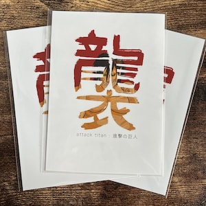 以下が含まれることがあります： 赤と金色の日本の文字デザインが施された3枚の白いカード。文字の下には「attack titan: 進撃の巨人」と書かれています。カードは透明なプラスチックの袖に入っており、木製の表面に置かれています。