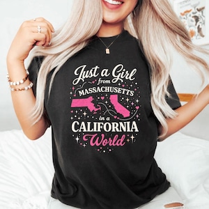 Puede incluir: Camiseta negra con el texto "Just a Girl from Massachusetts in a California World". El diseño incluye contornos rosas de los estados de Massachusetts y California, con estrellas y corazones decorativos.