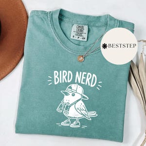 May include: A teal Comfort Colors t-shirt with the words "BIRD NERD" and a cartoon bird wearing a hat and holding binoculars. A silver necklace with a small pendant is also visible.