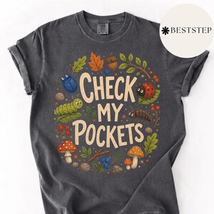 May include: Charcoal grey t-shirt featuring the phrase "CHECK MY POCKETS" in a playful font, encircled by drawings of insects, mushrooms, leaves, and berries. The design includes a blue beetle, a ladybug, and a green caterpillar.