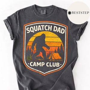 Può includere: T-shirt grigio scuro con una grafica raffigurante una silhouette di Bigfoot davanti a un tramonto e una tenda. Il testo recita "Squatch Dad Camp Club" in un design a scudo. La maglietta ha le maniche arrotolate.