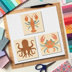 May include: Three square art prints featuring sea creatures: a lobster, an octopus, and a crab. The lobster is orange with blue and yellow accents. The octopus is brown. The crab is orange and blue. The prints are on a cream background.