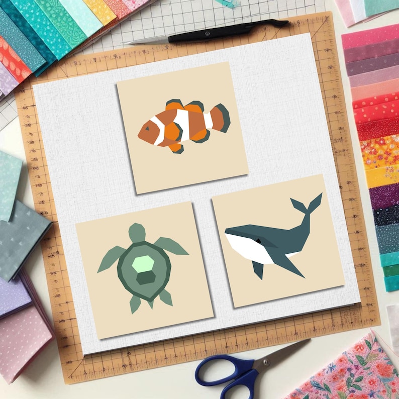 May include: Three square art prints with geometric designs of a clownfish, a sea turtle, and a whale. The prints are on a neutral background, with fabric scraps and sewing tools visible. The prints are likely for a craft project.