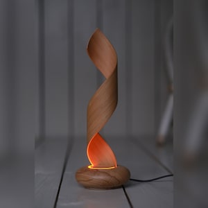 May include: A wooden table lamp with a unique, flowing design. The lamp features a spiraling, ribbon-like shade made of light-colored wood. The base is a rounded shape, and the lamp emits a warm, orange glow. The lamp is approximately 12 inches tall.