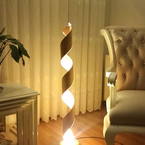 May include: A tall floor lamp featuring a spiraling, ribbon-like structure in a light tan color. The lamp emits a warm, bright glow from within the twists of its vertical frame, which is anchored by a circular base resting on a wooden floor.