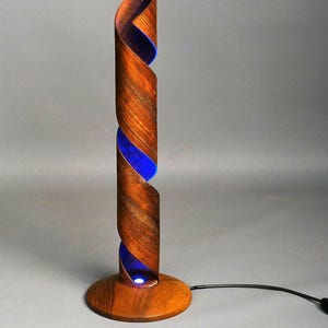 May include: A tall, cylindrical table lamp featuring a spiraling brown wood grain structure that reveals a vibrant blue interior light. The lamp stands on a circular wooden base and includes a black power cord extending from the side of the base.