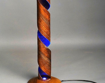 May include: A tall, cylindrical table lamp featuring a spiraling brown wood grain structure that reveals a vibrant blue interior light. The lamp stands on a circular wooden base and includes a black power cord extending from the side of the base.