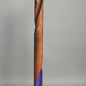 A wooden floor lamp with a unique design. The lamp is made of a single piece of wood that is twisted and shaped into a spiral. The lamp has a purple light that shines through the wood, creating a beautiful effect. The lamp is on a wooden base and has a black cord.
