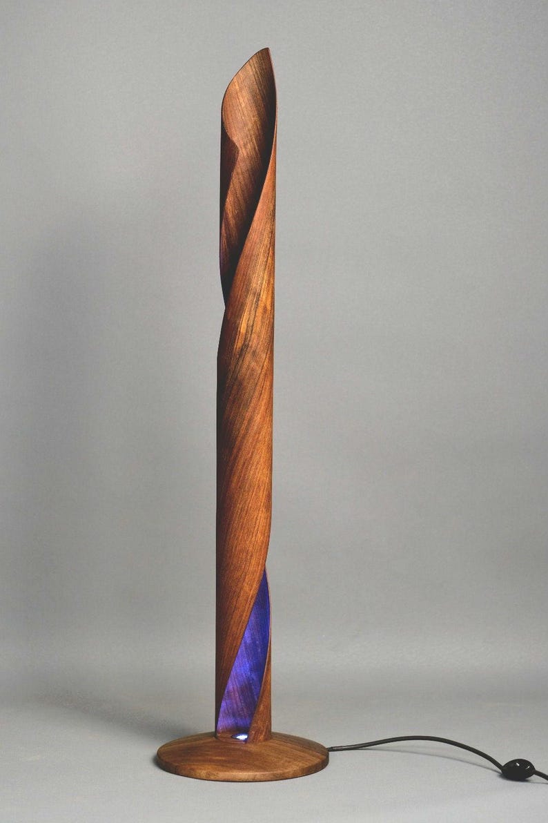 A wooden floor lamp with a unique design. The lamp is made of a single piece of wood that is twisted and shaped into a spiral. The lamp has a purple light that shines through the wood, creating a beautiful effect. The lamp is on a wooden base and has a black cord.