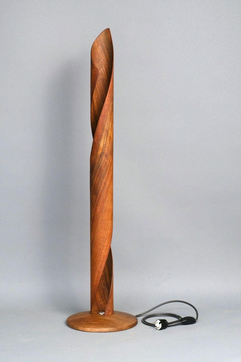 A wooden floor lamp with a twisted design. The lamp is made of dark brown wood and has a round base. The lamp has a black cord and plug.