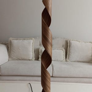 Op de afbeelding: Een houten tafellamp met een uniek, gedraaid ontwerp. De lampvoet is donkerbruin en rond. De lampenkap is gemaakt van dunne, lichtbruine houten stroken die met elkaar zijn gedraaid om een spiraalvorm te creëren.