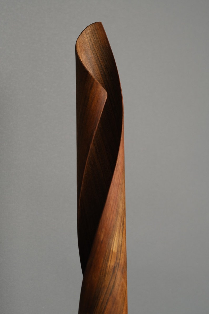 A wooden sculpture of a twisted, spiraling form. The sculpture is made of dark brown wood with a natural grain pattern. The sculpture is tall and slender, and it appears to be made of multiple pieces of wood that have been joined together.