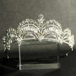 Può includere: Una tiara d'argento con una fila di strass trasparenti e un designo decorativo al centro. La tiara è su un supporto in vetro trasparente.