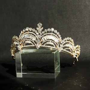 Può includere: Una tiara dorata con decorazioni in cristallo trasparente. La tiara è su un supporto in vetro trasparente.