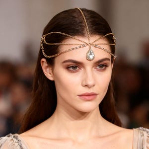 May include: A gold-toned headpiece with a delicate chain design. The headpiece features a central teardrop-shaped clear crystal and smaller crystals along the chain. The accessory is worn on a person with long, dark hair.