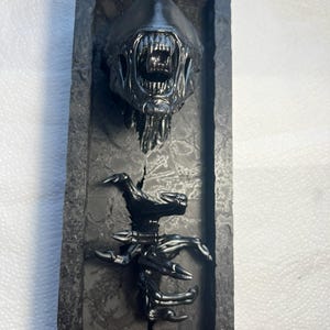 Op de afbeelding: Een zwart en grijs alien-buste met een open mond en scherpe tanden, gemonteerd in een zwart frame. De hand van de alien is ook zichtbaar, die uit het frame steekt.