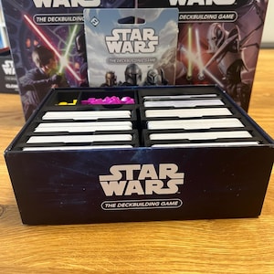 Pode incluir: Um conjunto de caixas do jogo de construção de baralhos Star Wars. O conjunto inclui uma caixa com o título do jogo, um pacote de cartas menor e duas caixas de jogo com ilustrações de personagens. A caixa do jogo é preta com texto branco e um gradiente azul.