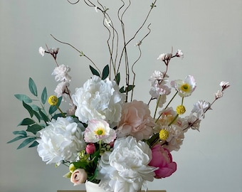 以下が含まれることがあります： 白い陶器の花瓶に、白い牡丹、ピンクのバラ、白とピンクの野花を組み合わせたフラワーアレンジメントが活けられています。アレンジメントは緑の葉と茶色の枝で飾られています。