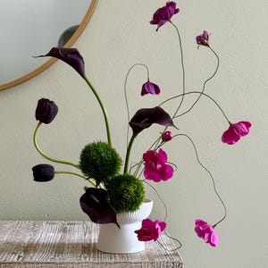May include: A floral arrangement featuring dark purple calla lilies, magenta flowers, and green spherical accents in a white vase. The arrangement is set against a neutral background, creating a striking contrast.