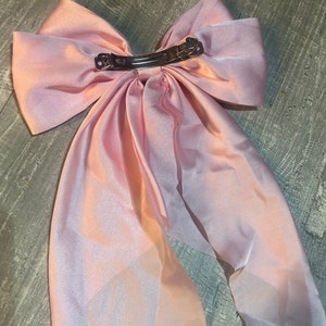 May include: A large, light pink satin hair bow with a silver metal clip. The bow is made of a soft, flowing fabric and has long, draping tails. The clip is attached to the back of the bow for easy wear.