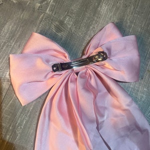 May include: A large, light pink satin hair bow with a silver metal clip. The bow is made of a soft, flowing fabric and is designed to be worn in the hair. The clip is marked "CLUB PARIS".