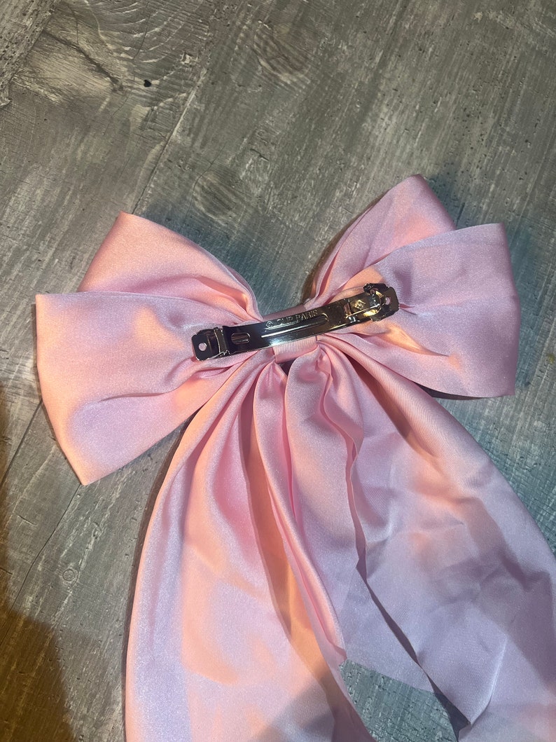 May include: A large, light pink satin hair bow with a silver metal clip. The bow is made of a soft, flowing fabric and is designed to be worn in the hair. The clip is marked "CLUB PARIS".