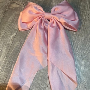 May include: A large, light pink satin bow with long, flowing tails. The bow is centered and has a classic shape, with a knot in the middle. The fabric has a subtle sheen, and the overall effect is elegant and feminine.