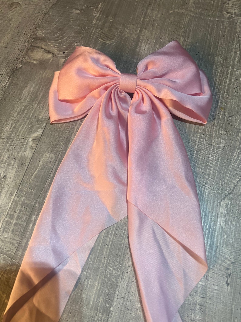 May include: A large, light pink satin bow with long, flowing tails. The bow is centered and has a classic shape, with a knot in the middle. The fabric has a subtle sheen, and the overall effect is elegant and feminine.