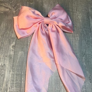 May include: A large, light pink satin bow with long, flowing tails. The bow is tied in the center and has a full, voluminous shape. The fabric has a slight sheen, and the bow is set against a gray wooden surface.