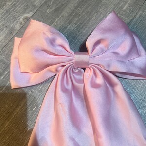May include: A large, light pink satin bow. The bow is tied in the center with long, flowing tails. The fabric has a subtle sheen, and the bow is likely used as a hair accessory or decorative element.