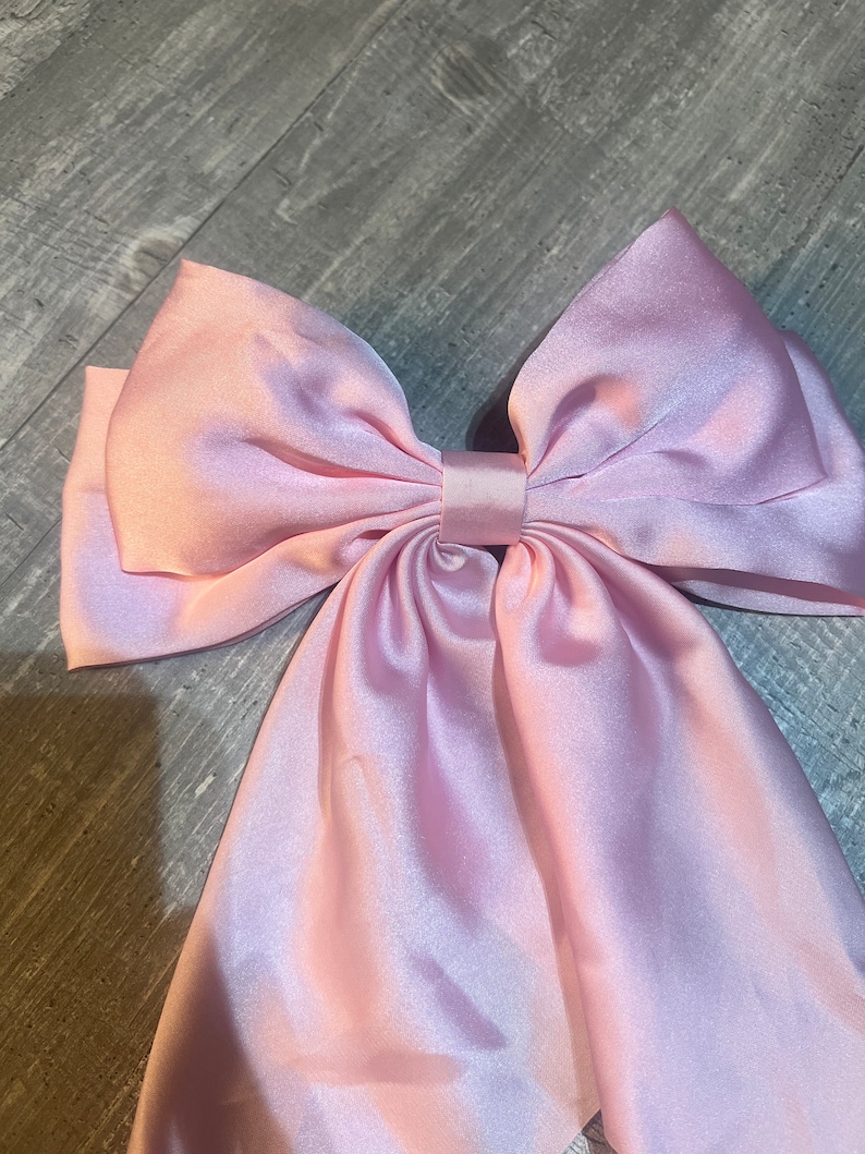 May include: A large, light pink satin bow. The bow is tied in the center with long, flowing tails. The fabric has a subtle sheen, and the bow is likely used as a hair accessory or decorative element.