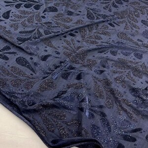 May include: Navy blue fabric with a shimmering, leaf-like pattern. The design features dark blue and silver glitter accents, creating a textured and elegant appearance. The fabric is folded, showing its drape and sheen.
