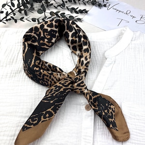 Può includere: Una sciarpa di seta con stampa leopardata nera, marrone e beige. La sciarpa è annodata e appoggiata su una camicia bianca. La sciarpa quadrata è realizzata in un tessuto morbido e fluido.