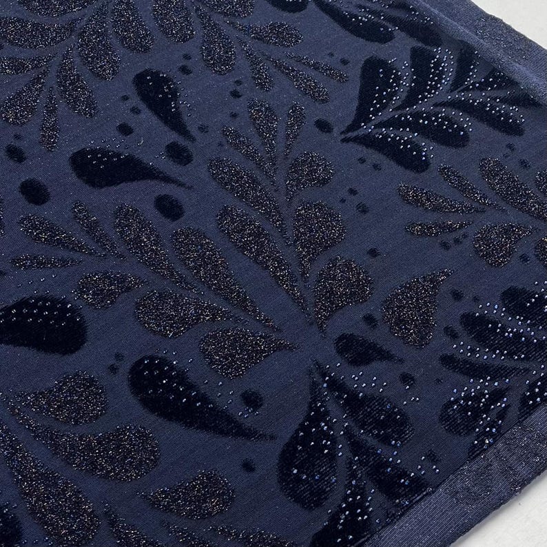 May include: A close-up shot of a navy blue fabric with a repeating pattern of stylized leaves and teardrop shapes. The design features a velvet texture with sparkling glitter accents, creating a luxurious appearance. The fabric has a subtle sheen.