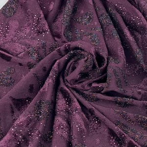 May include: Close-up of a deep purple fabric with a velvet texture. The fabric has a pattern of dark, glittery shapes and scattered iridescent sequins. The material is draped, showing folds and shadows.