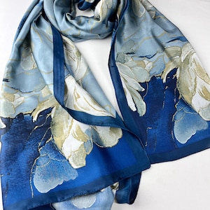 Op de afbeelding: Blauwe zijden sjaal met een bloemenpatroon in wit en goud. De sjaal heeft een donkerblauwe rand en is gedrapeerd. De stof lijkt zacht en vloeiend. Het ontwerp is elegant en geschikt voor diverse gelegenheden.