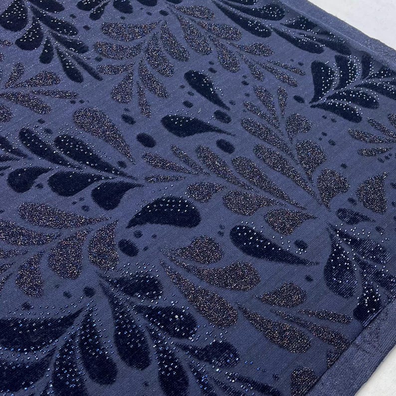May include: A close-up shot of a navy blue fabric featuring a repeating pattern of stylized leaf shapes. The leaves are a darker navy with a velvet texture and are embellished with tiny, sparkling silver dots. The background is a solid navy blue.